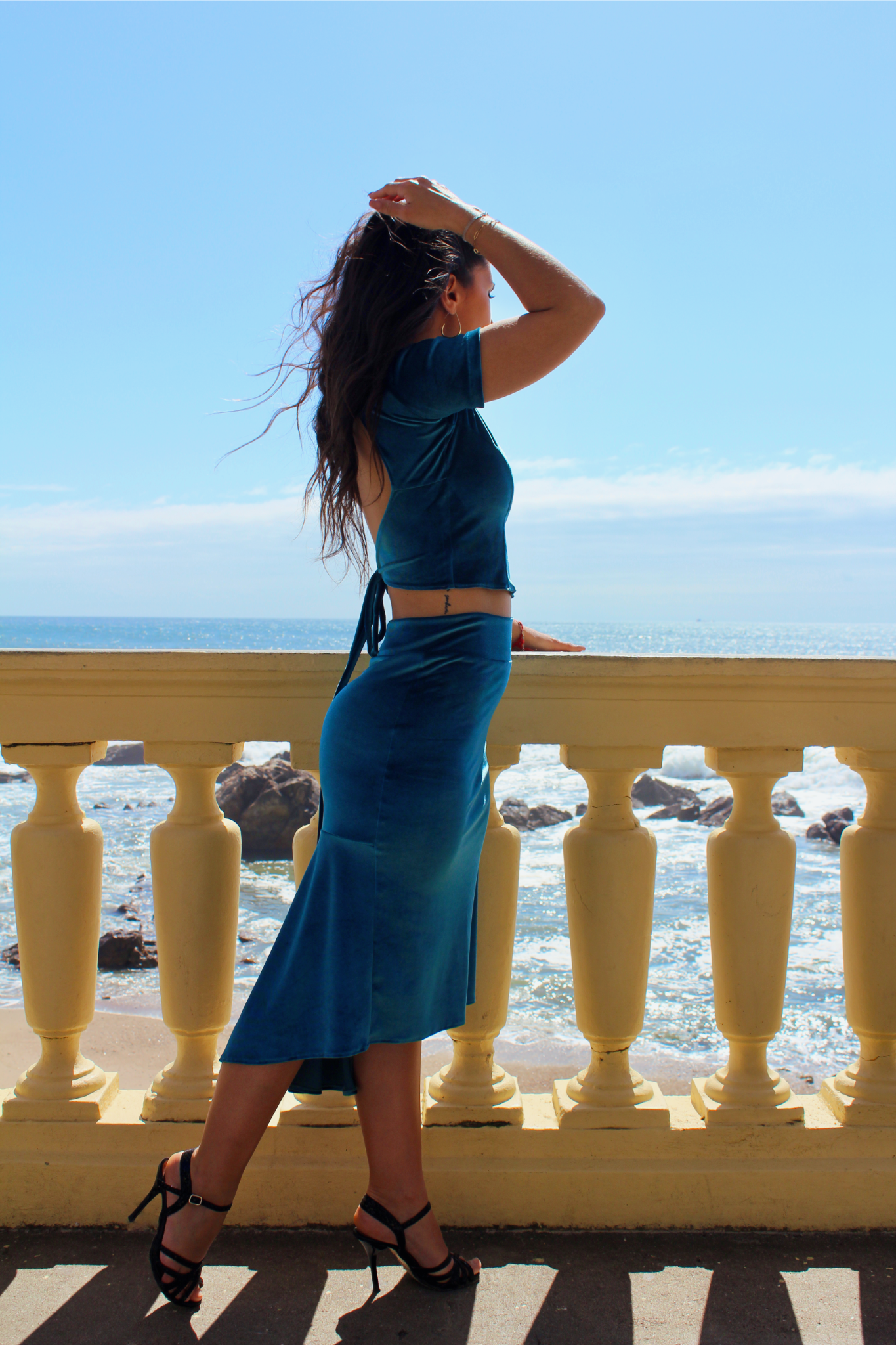 Side view of a tango dancer wearing the Afrodite Turquoise blue velvet set by De Floreo, looking at the ocean.